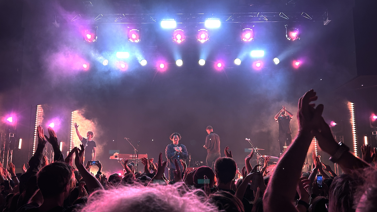 Indie rock band The Temper Trap performing live on stage in front of a cheering crowd under bright pink and blue concert lights.